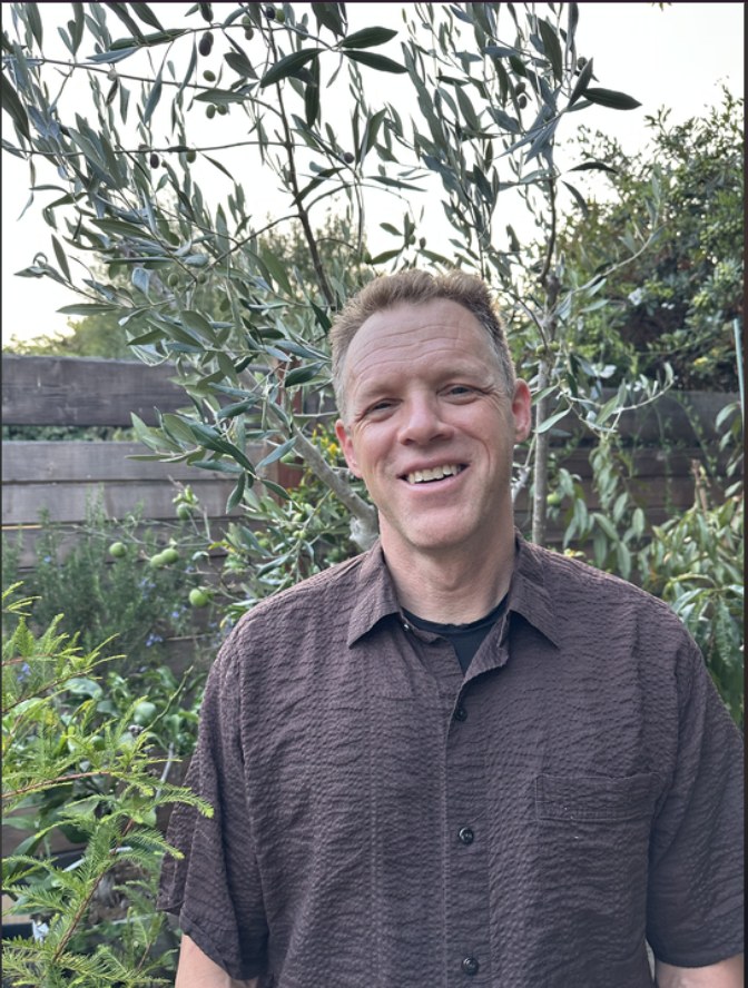 Daniel Sapoznick, LCSW, standing outdoors beside an olive tree in a garden